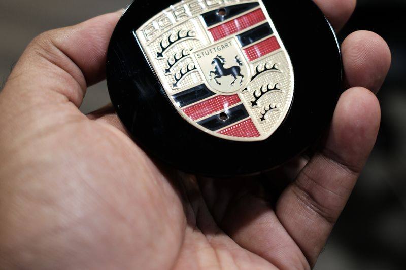 A hand holding a black wheel centre cap carrying the Porsche crest.