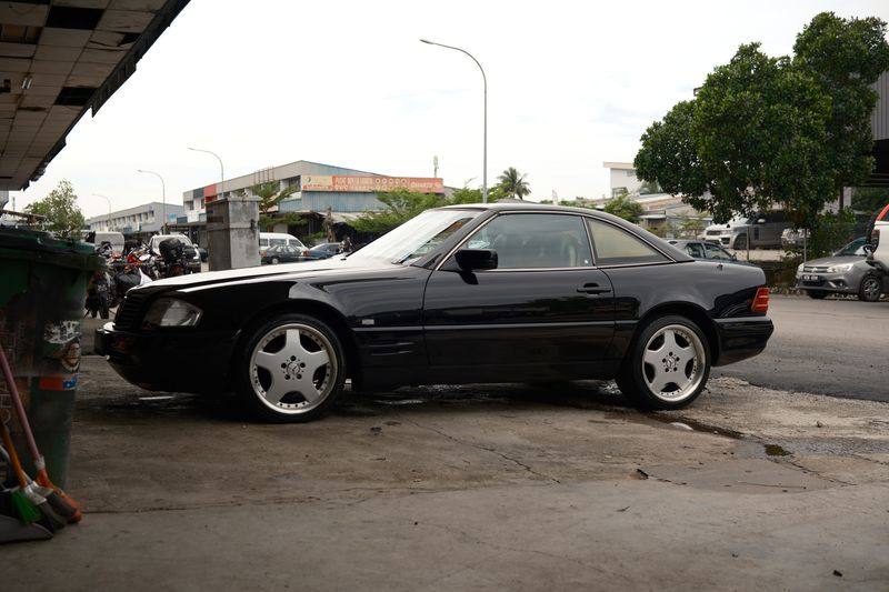 A black Mercedes-Benz R129 SL in profile with its hardtop fitted, parked on a workshop apron.