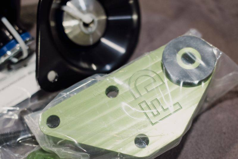 A billet engine mount with a polished centre next to a green anodised bracket in a bag.